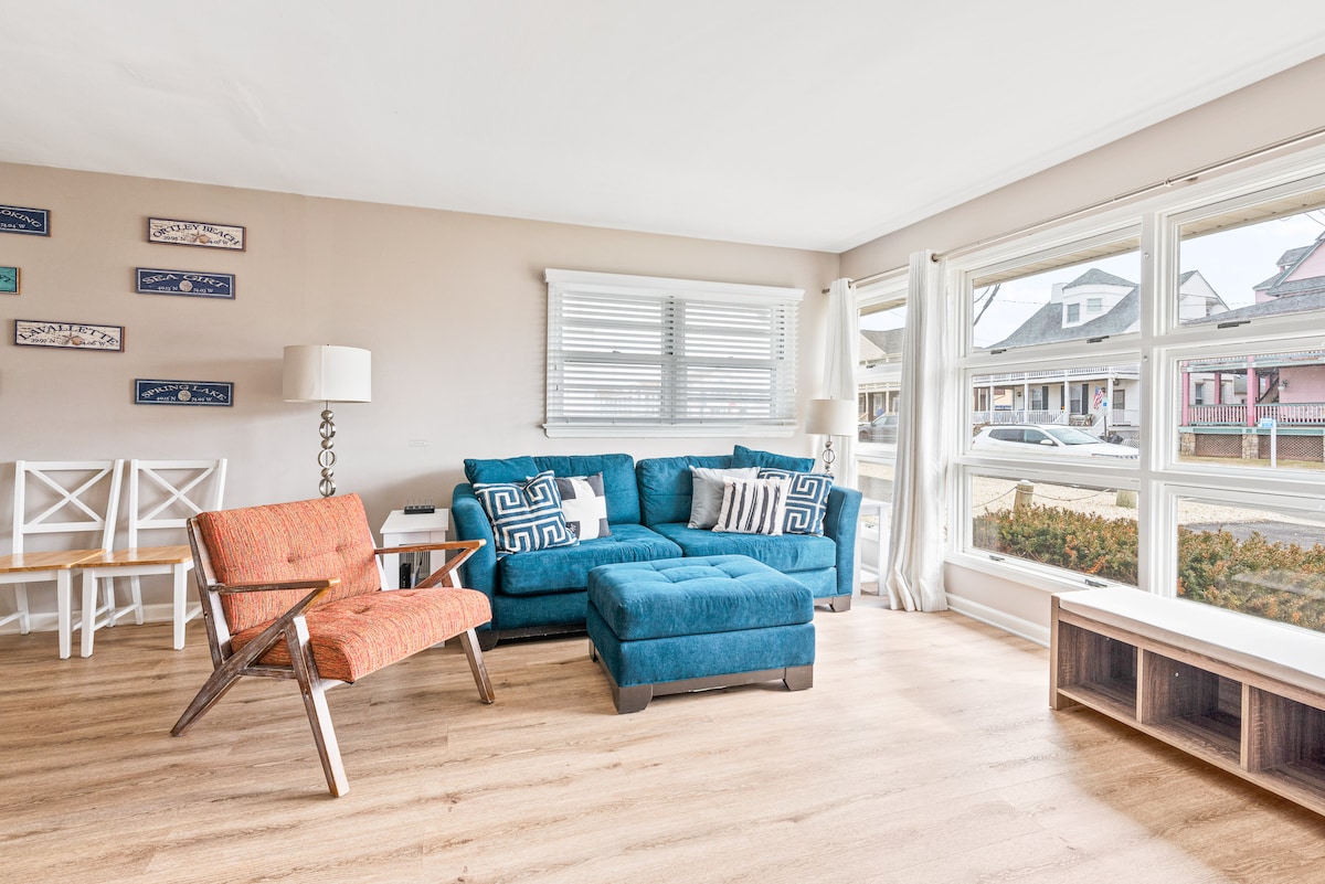 A bright living area features a teal sectional sofa paired with an orange accent chair. Large windows allow natural light to fill the space, while light wood flooring complements the decor. A lamp and a media unit provide additional functionality.