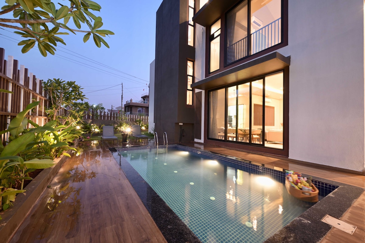 A modern villa exterior features a private pool with clear water, bordered by wooden decking and tropical plants. Large glass doors offer views into the interior space, which is softly illuminated as dusk settles.