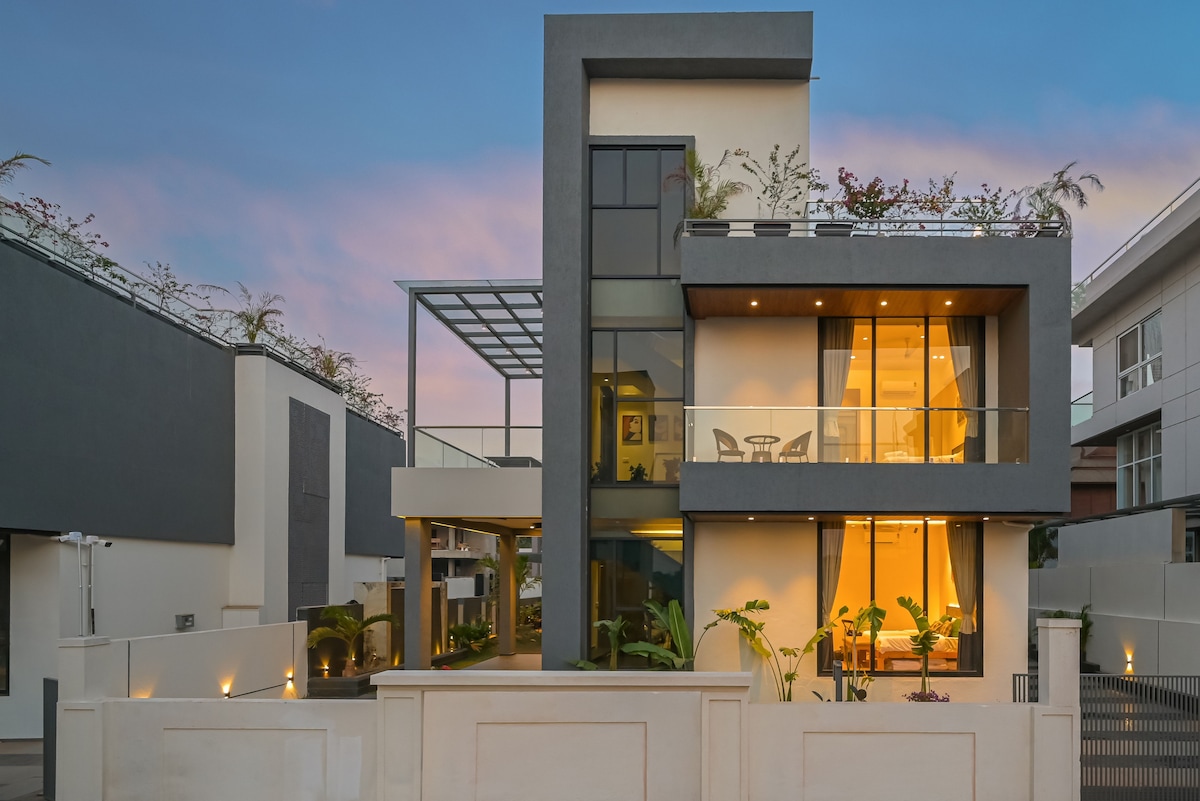 The modern villa's exterior features a two-story design with large glass windows allowing natural light to fill the space. Potted plants and greenery are visible around the entrance, enhancing the welcoming appearance. A covered terrace area is seen at the upper level, overlooking the surroundings.