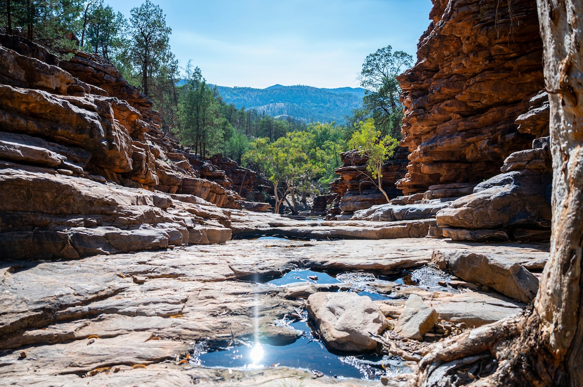 A serene landscape features rugged rock formations lining a narrow waterway, reflecting sunlight. Lush greenery is visible among the rocky outcrops, with distant mountain ranges forming the backdrop against a clear blue sky.