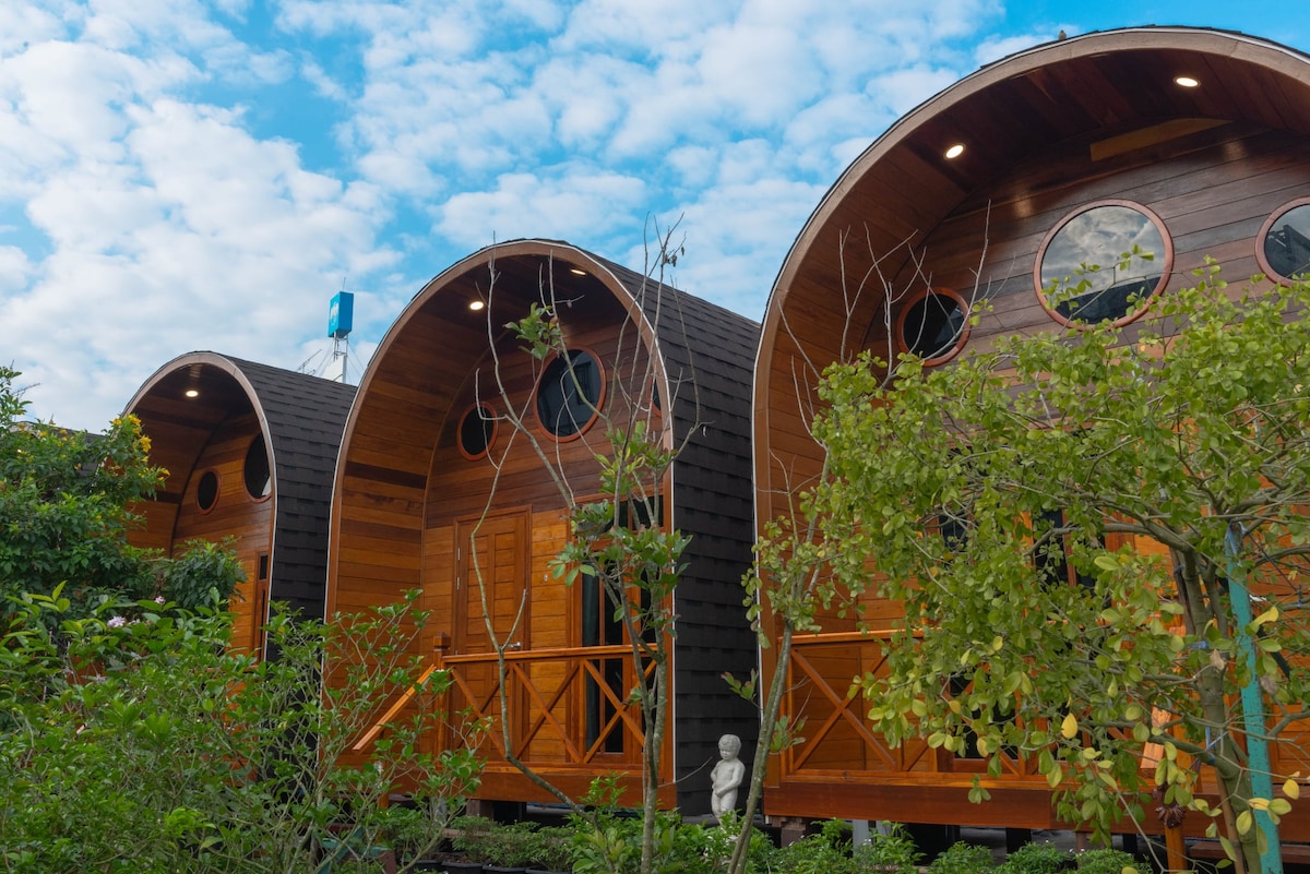 Three wooden cabins feature rounded roofs and circular windows, nestled among greenery. The natural wood finishes are highlighted by soft lighting, and a small statue can be seen in the foreground, contributing to a peaceful outdoor atmosphere.
