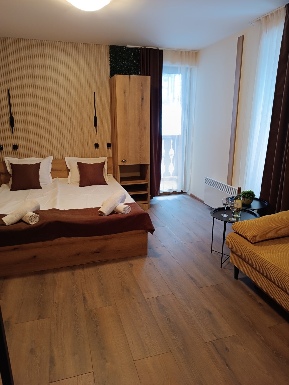 A spacious sleeping area features a comfortable bed with decorative pillows and neatly arranged towels. Natural light filters through sheer curtains covering the window. A cozy seating area includes a small table and a charming armchair, enhancing the room's inviting layout.