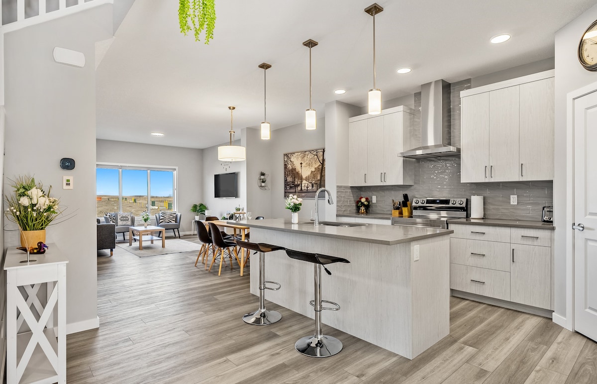 An open-concept kitchen and living area showcases modern design with a large island and sleek cabinetry. Pendant lights hang above the island, and bar stools offer seating. A living space is visible in the background, featuring comfortable seating and a media setup, with natural light streaming in through sliding glass doors.