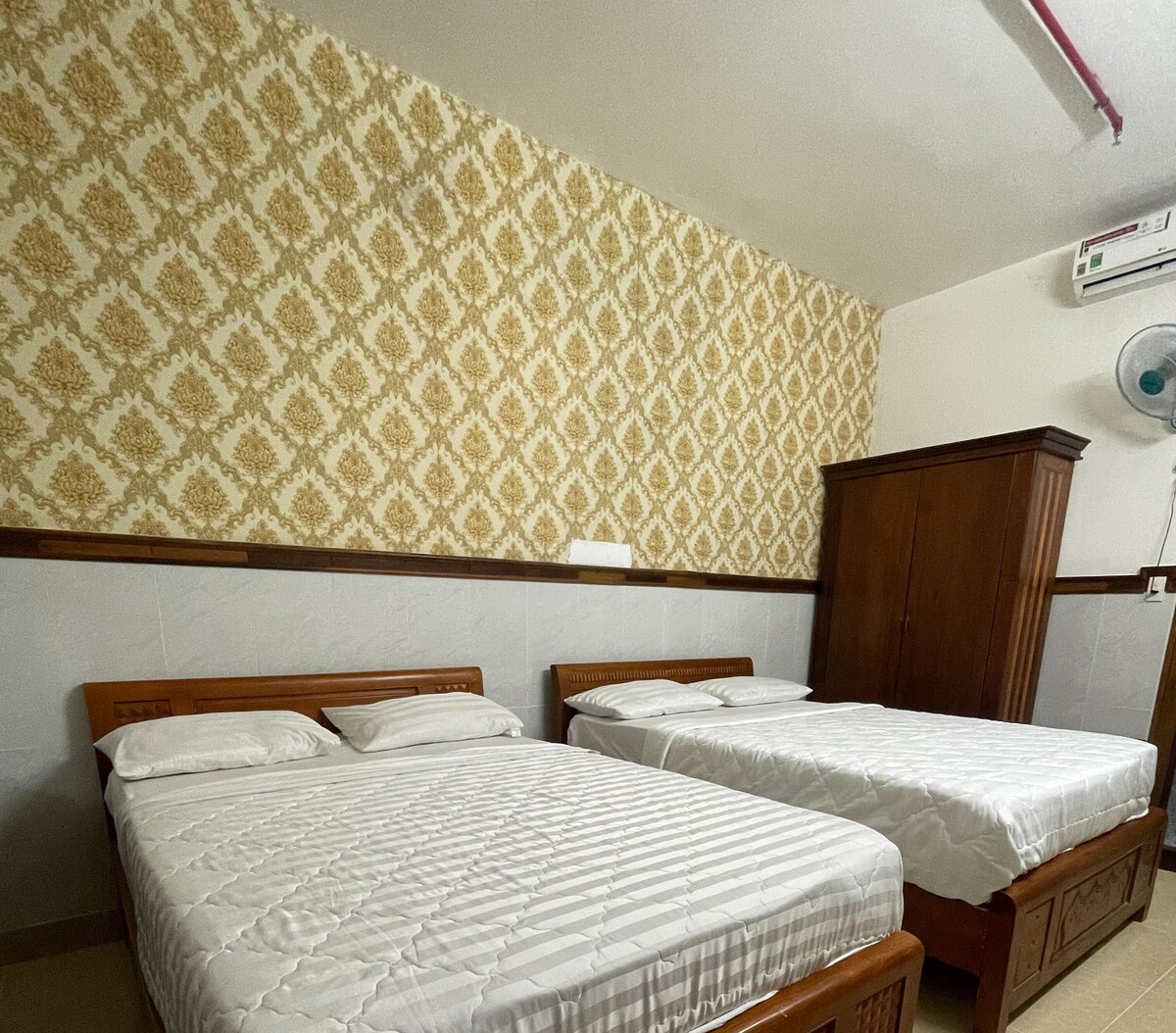 The sleeping area features two spacious beds, dressed in white linens. A decorative gold-patterned wallpaper adorns the walls, complemented by a wooden wardrobe. An air conditioning unit and a ceiling fan provide climate control, ensuring comfort throughout the stay.