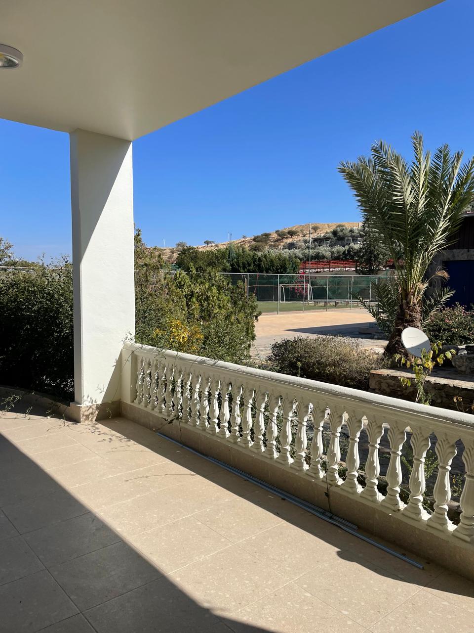 A patio area is framed by a white balustrade, leading to a view of greenery and a distant hillside. Clear blue skies enhance the open feel, while the surrounding plants offer a natural touch to the outdoor space.