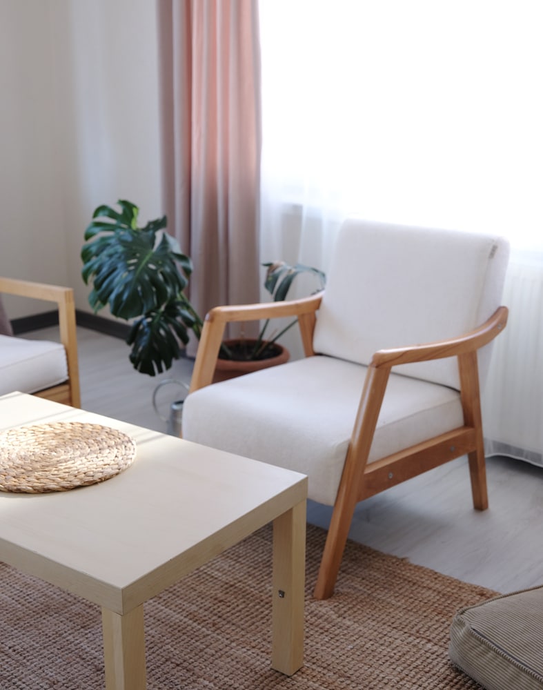 Light fills a relaxing living space, featuring a simple wooden coffee table with a woven placemat. A pair of comfortable armchairs with light upholstery are positioned around the table, complemented by a potted plant nearby. Soft curtains add warmth to the environment.