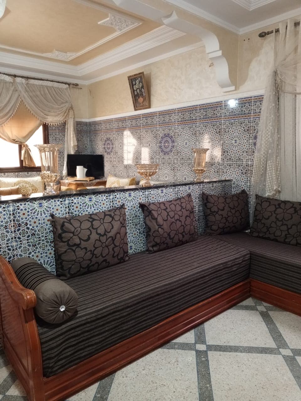 A comfortable seating area is provided, featuring a low wooden platform adorned with dark cushions and decorative pillows. A detailed tile backsplash adds vibrancy, while warm sunlight filters through sheer curtains. A side table displays an arrangement of decorative items.