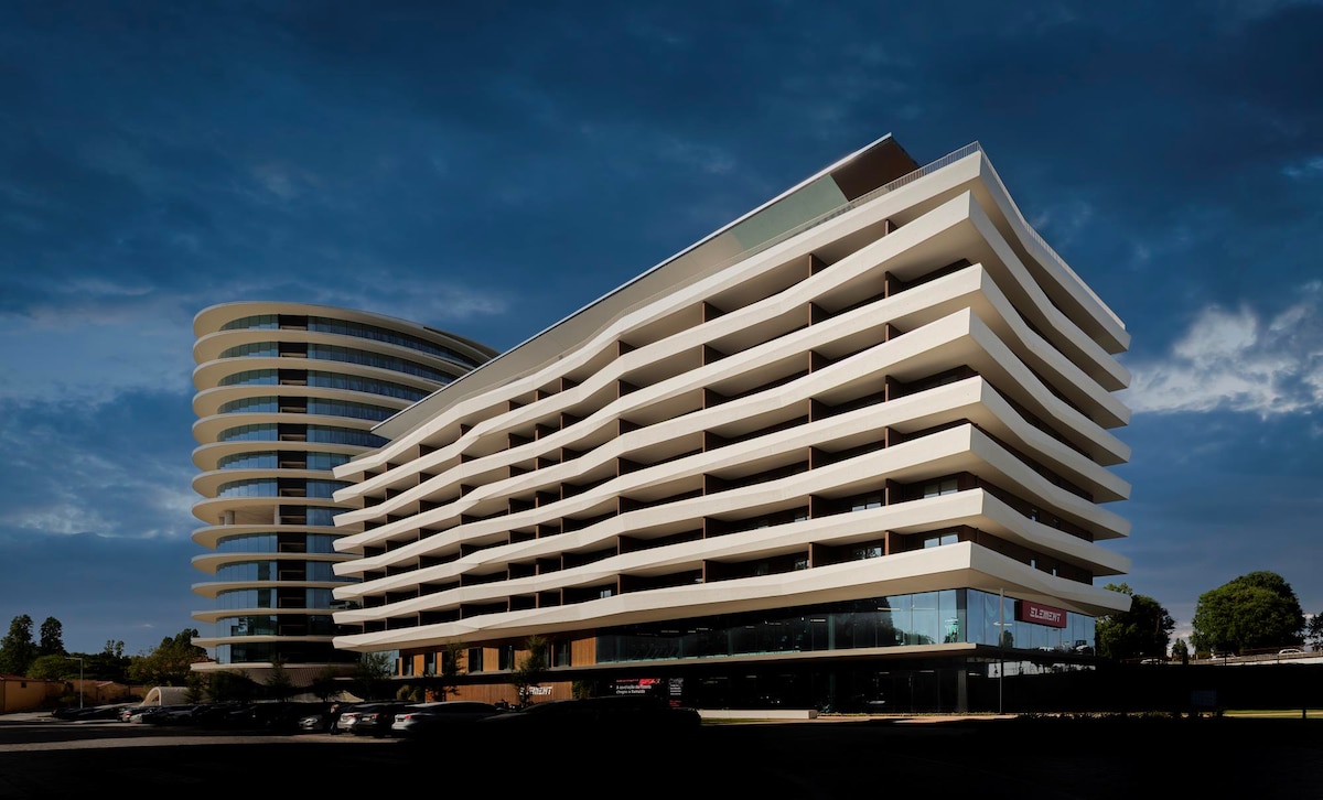 A modern building structure is presented with multiple levels, featuring an array of balconies and large windows. The design combines curved and linear elements, with a glass-enclosed ground level that provides visibility into the lobby area. The surrounding area appears to be landscaped.