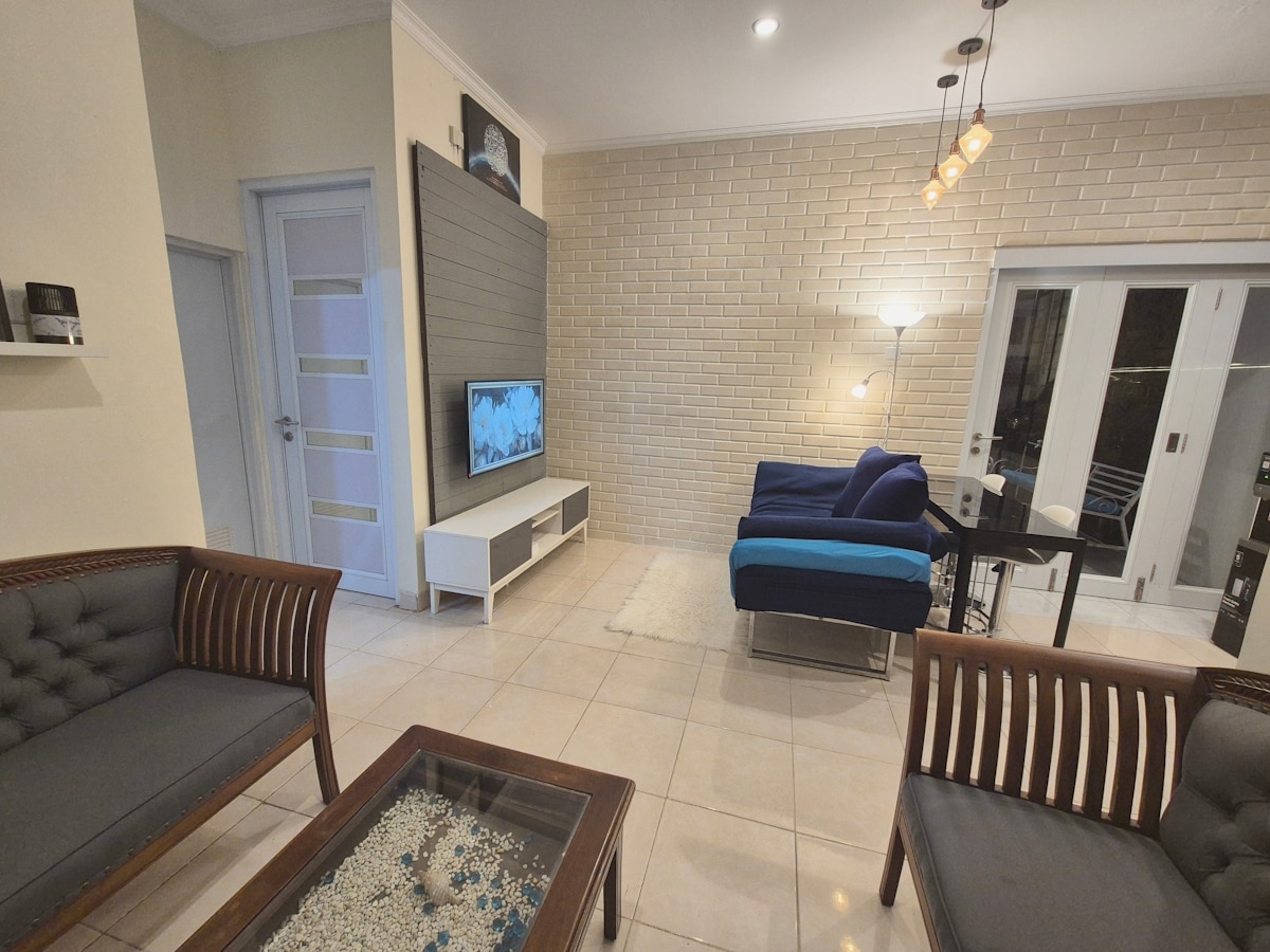 A living area features two upholstered chairs facing a coffee table with decorative items. A sectional sofa in blue is positioned beside a TV unit. Bright tiles cover the floor, and natural light is allowed through glass doors leading to an outdoor space.