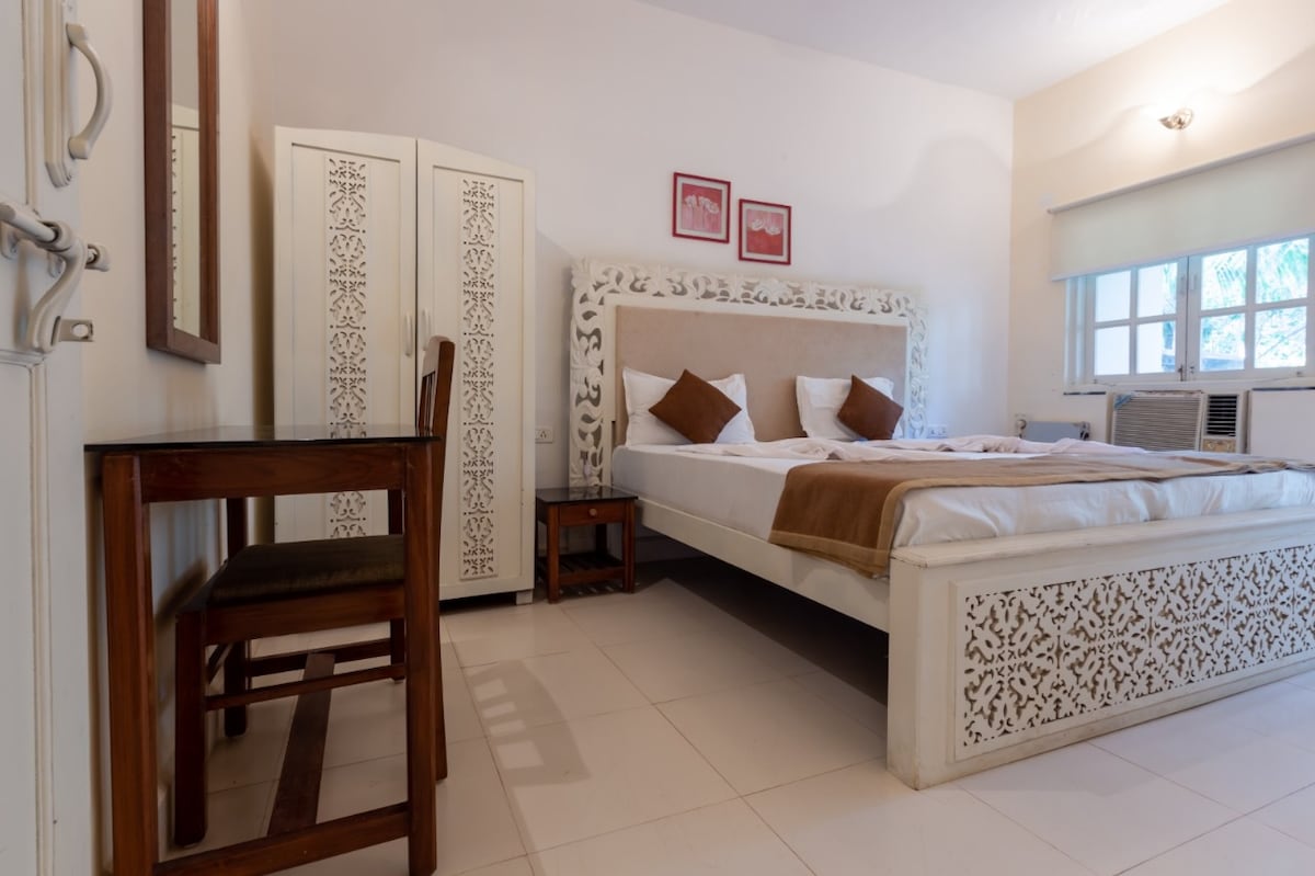 A spacious bedroom features a large, ornate bed with a decorative headboard and two accent pillows. A wooden desk and chair provide a work area. Natural light enters through a window with sheer curtains, and an air conditioning unit is visible in the corner.