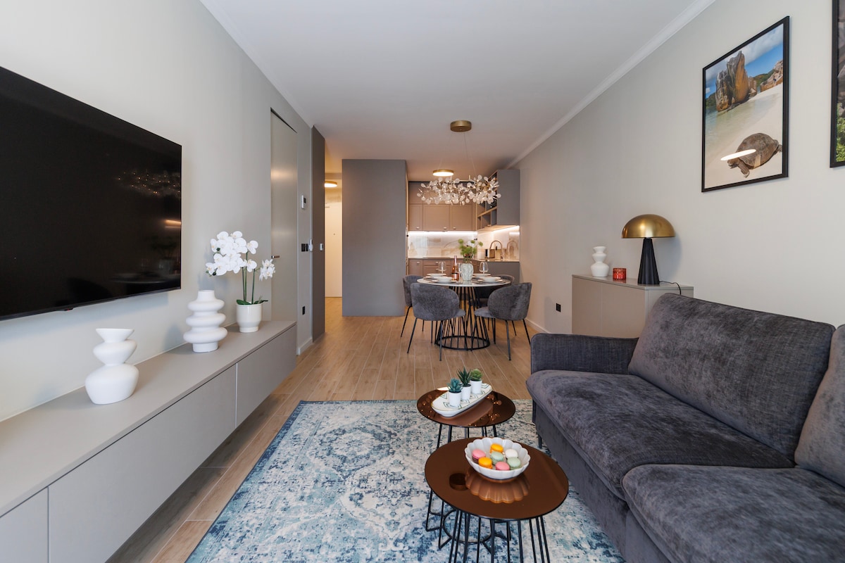 A contemporary living area features a plush dark sofa facing a wall-mounted television. A stylish coffee table displays decorative items. In the background, a dining space with four upholstered chairs is visible, alongside a modern kitchen illuminated by pendant lights.
