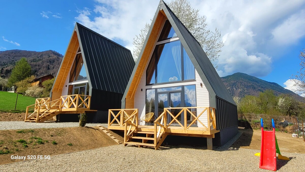 Two A-frame cabins are presented, featuring large windows that allow for ample natural light. Each cabin has a wooden deck with railing, and wooden lounge chairs are positioned for outdoor seating. A playground with a slide is visible in the foreground, surrounded by green grass.