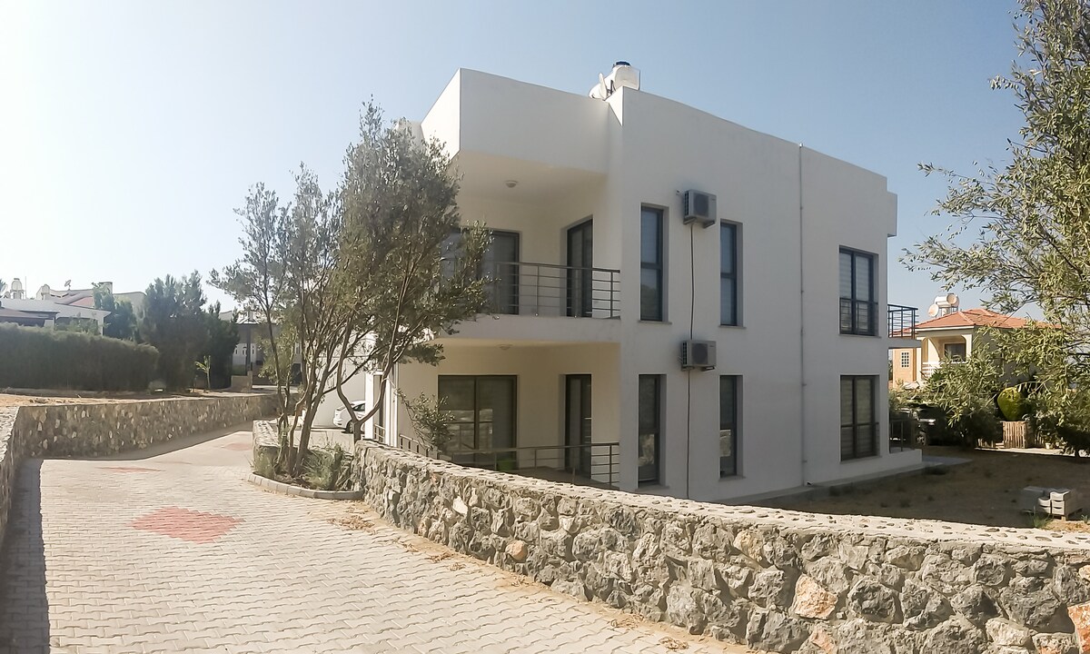 A modern two-story building is shown, featuring large windows and a sleek, white exterior. A stone pathway leads to the entrance, while manicured trees and low shrubs provide greenery around the property. Air conditioning units are visible on the façade.