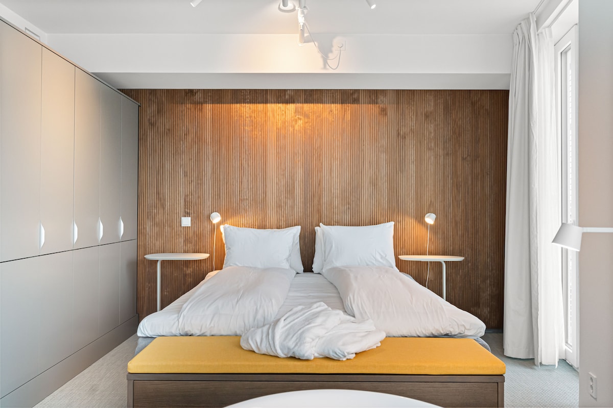 A well-appointed bedroom features a light wood accent wall behind two neatly made beds with white bedding. Complimentary nightstands with reading lamps are positioned on either side. Natural light enters through sheer curtains, providing a calm and airy feel to the space.