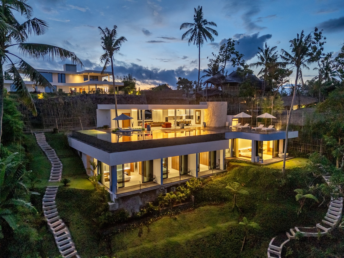 A modern villa is set amidst lush gardens, featuring large glass doors that open to a spacious terrace. The structure has a unique design, with palm trees framing the serene setting. Soft lighting highlights the open living spaces as twilight descends.