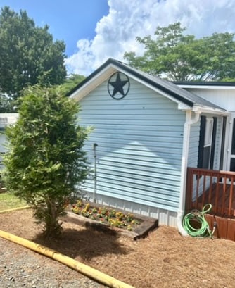 The exterior of the home is showcased, featuring light blue siding and a star decoration above the roofline. A small tree and flower bed with blooms are positioned in front, contributing to the surroundings. A wooden deck and a portion of the gravel driveway are visible.