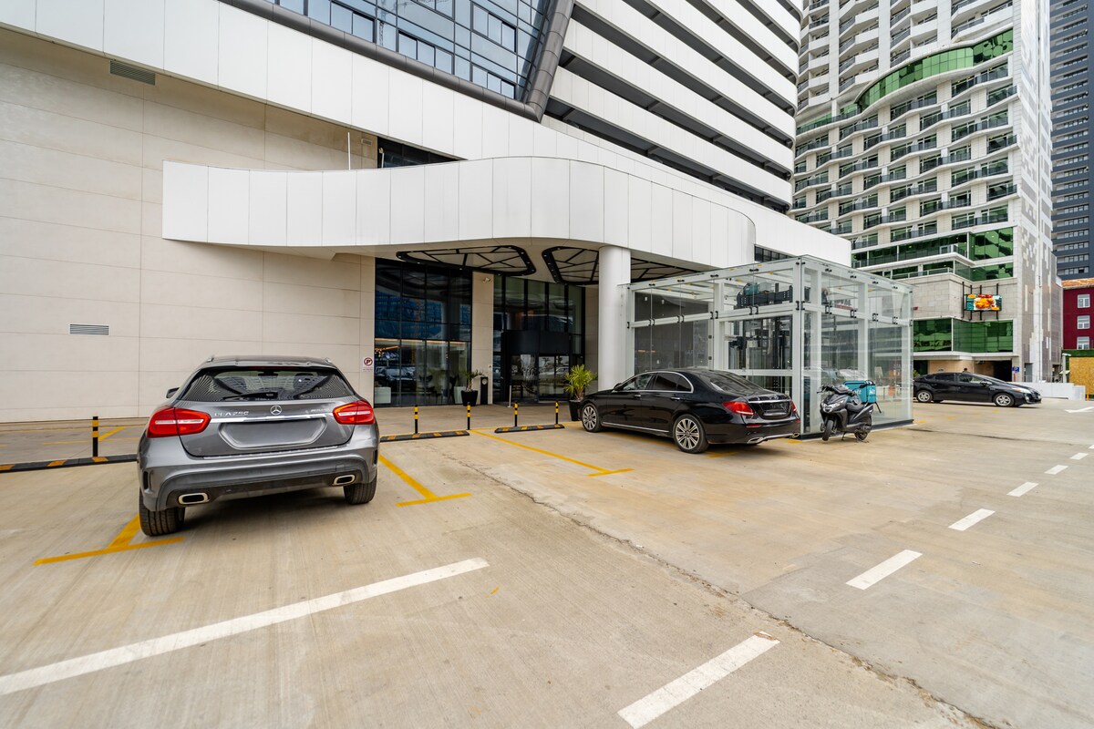 The image captures a modern building entrance with a sleek facade. Several parked vehicles are visible in the foreground, along with a glass structure that may serve as a lobby or entrance area. Clear lines and geometric shapes emphasize the contemporary design.