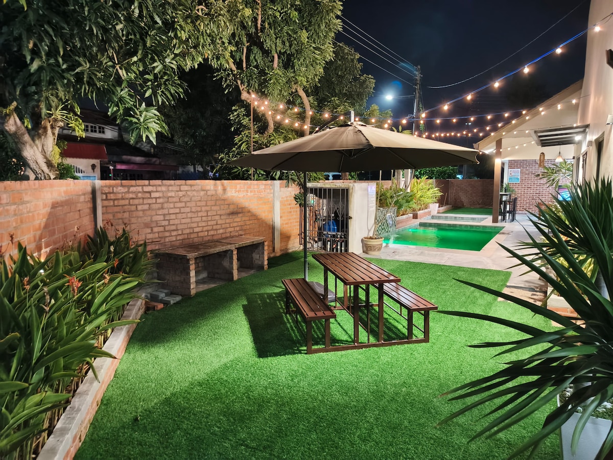 An outdoor space is illuminated softly by string lights, providing a cozy atmosphere. A wooden table and bench set is situated on lush green artificial grass, with manicured plants surrounding the area. In the background, a private pool is visible, reflecting the nighttime ambiance.