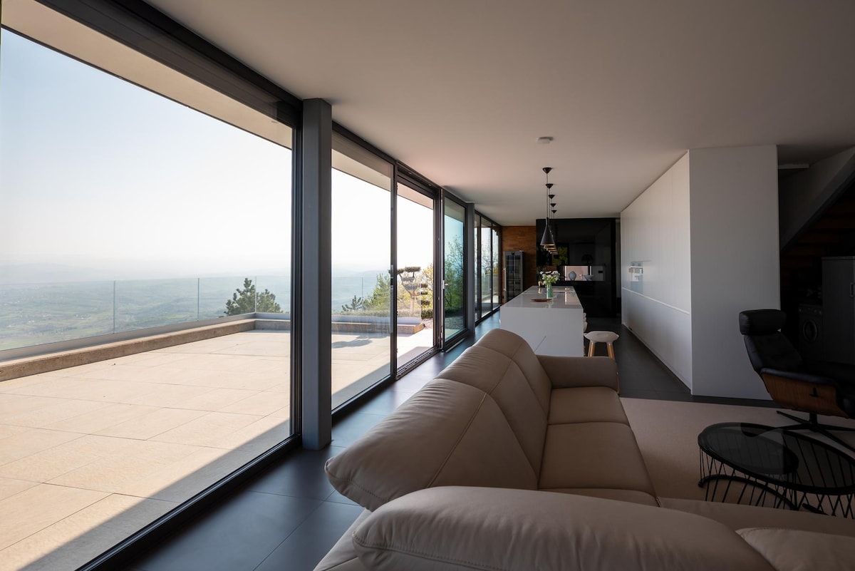 A spacious living area features floor-to-ceiling glass walls, allowing for an abundance of natural light and panoramic views of the surrounding landscape. A plush sectional sofa and a sleek dining area create a comfortable atmosphere, while a modern kitchen island enhances the contemporary design.