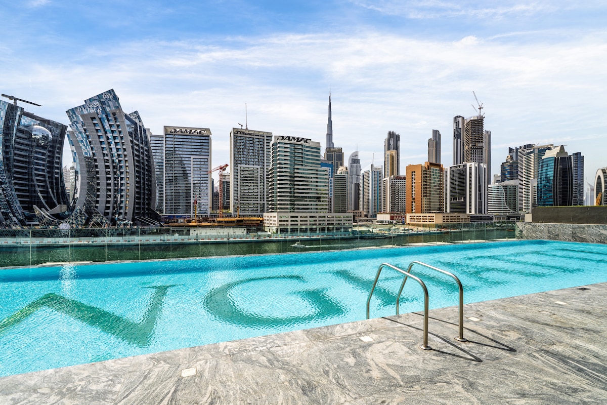 A serene infinity pool reflects the city skyline, featuring modern architecture and the iconic Burj Khalifa. The pool's edge is lined with sleek stone surfaces, and a set of metallic steps leads into the clear water, inviting relaxation and enjoyment of the city views.