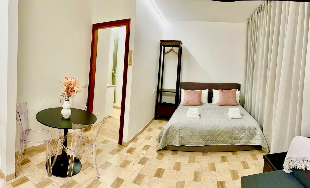 A spacious room features a large bed with decorative pillows and a quilted bedspread. A dining area includes a round table and transparent chairs. A wardrobe stands nearby, and light fabrics drape over the window, allowing natural light to enter, creating a relaxed atmosphere.