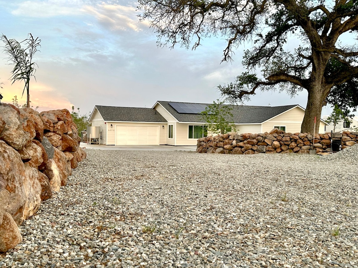 A spacious gravel driveway leads up to a single-story home, characterized by a simple exterior and a prominent oak tree nearby. Stone walls are visible in the foreground, framing the scene and enhancing the rural ambiance. Solar panels are installed on the roof.