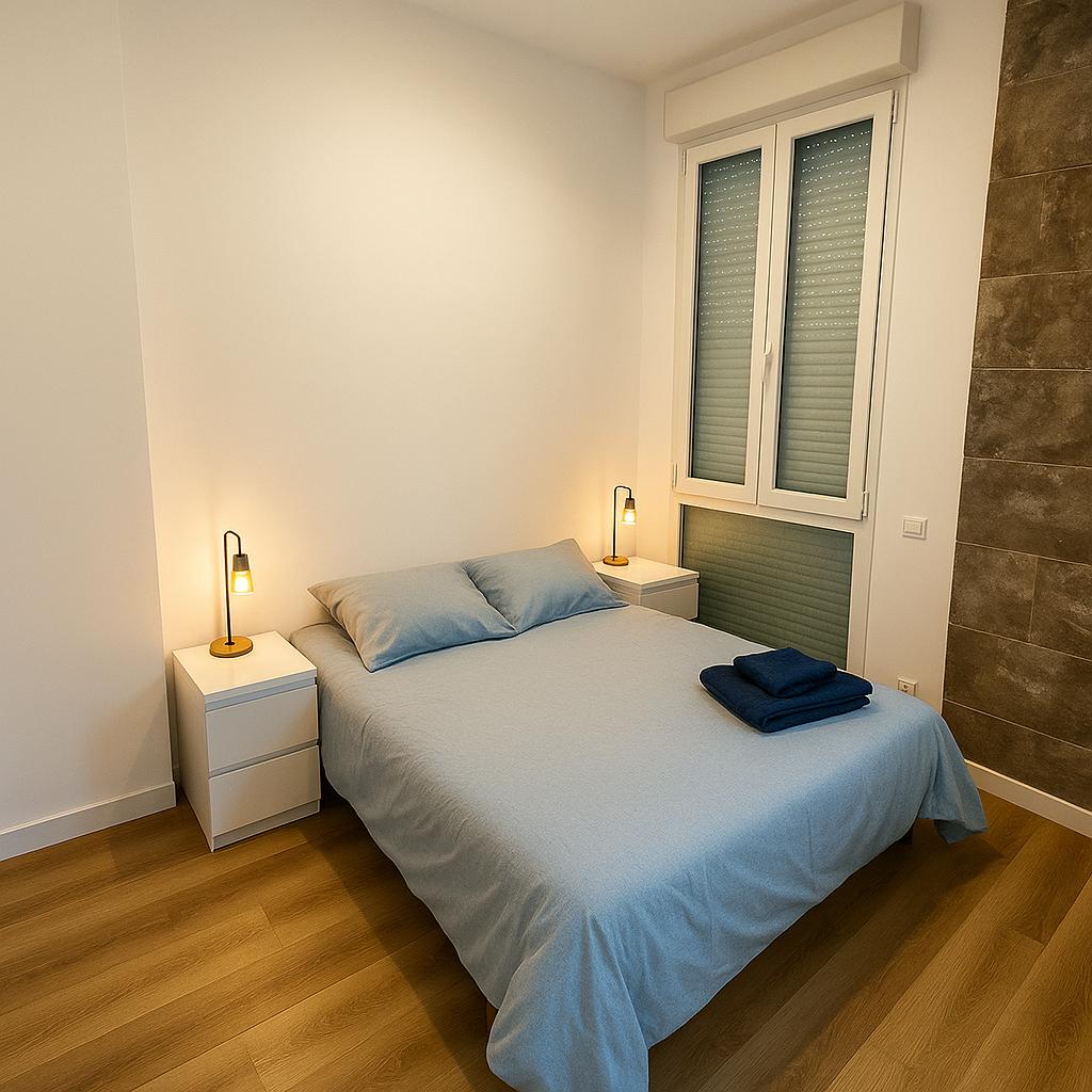 A bright, modern bedroom is presented with a spacious bed covered in a light blue blanket. Two bedside tables with stylish lamps flank the bed, while a large window allows natural light to fill the space. Light wood flooring adds warmth to the room.