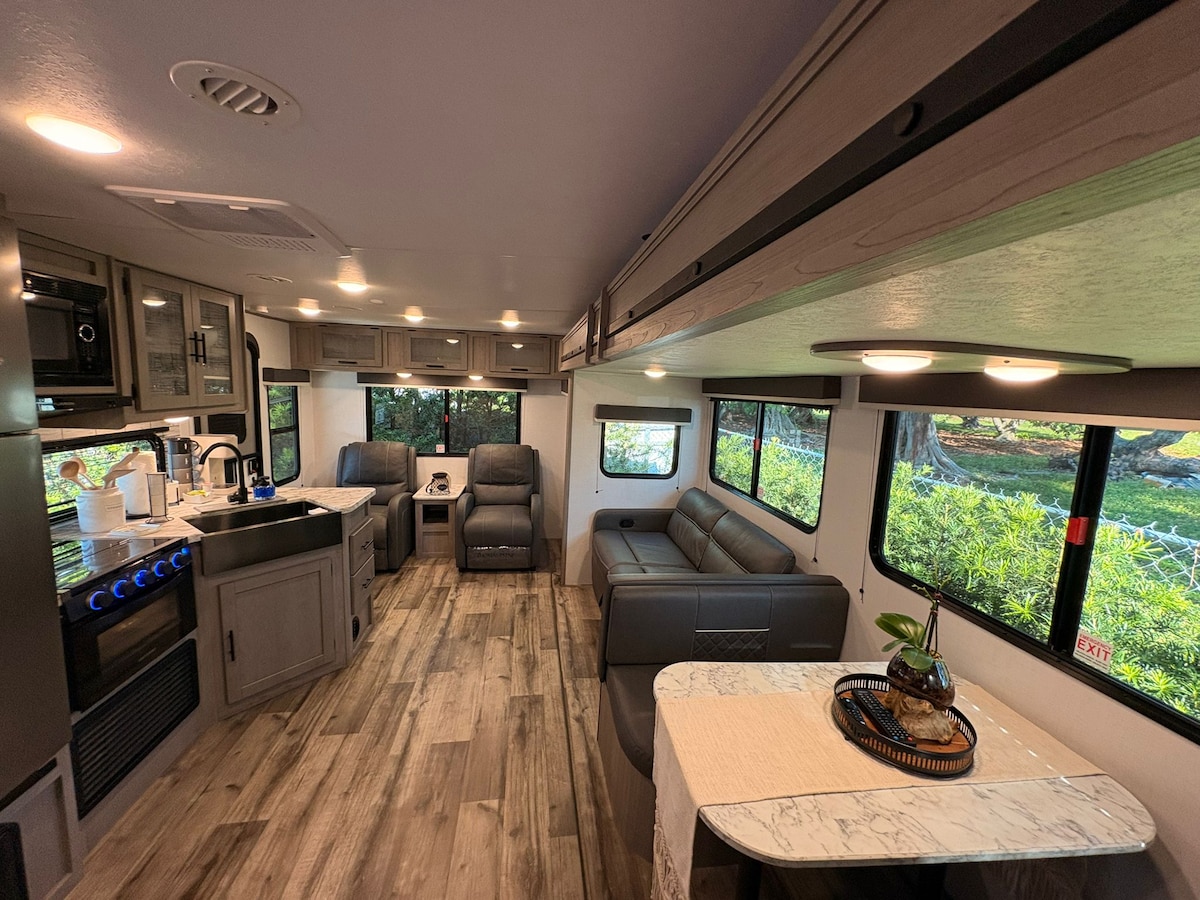 An inviting interior of the RV is shown, featuring a spacious seating area with a gray sectional sofa. The kitchenette is equipped with modern appliances. Large windows allow natural light to fill the space, while wood-like flooring enhances the cozy atmosphere. A dining table is also visible.