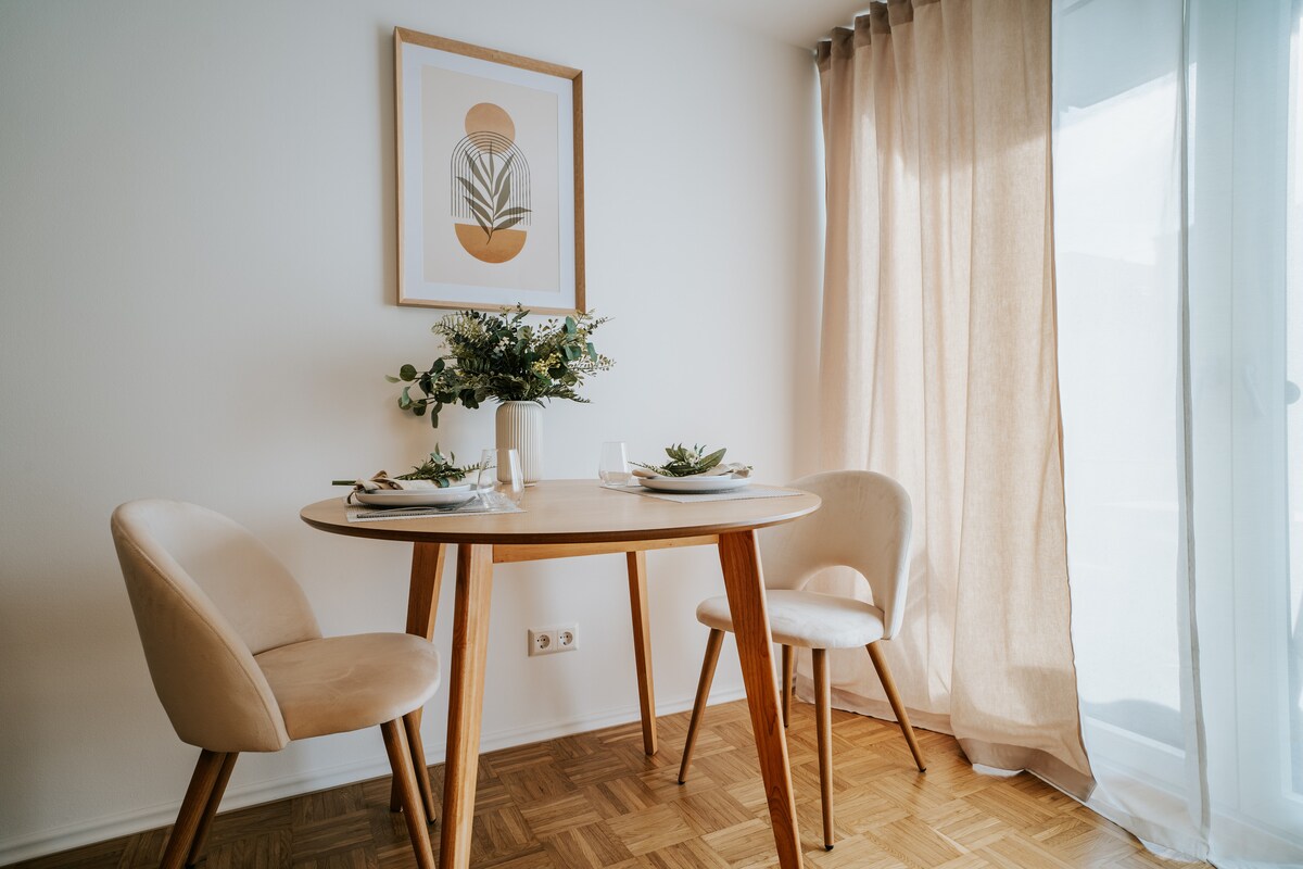 A light and airy dining space features a round wooden table set for two, complemented by soft beige chairs. A minimalist wall art piece decorates the wall above, while sheer curtains softly diffuse natural light from the adjacent window.