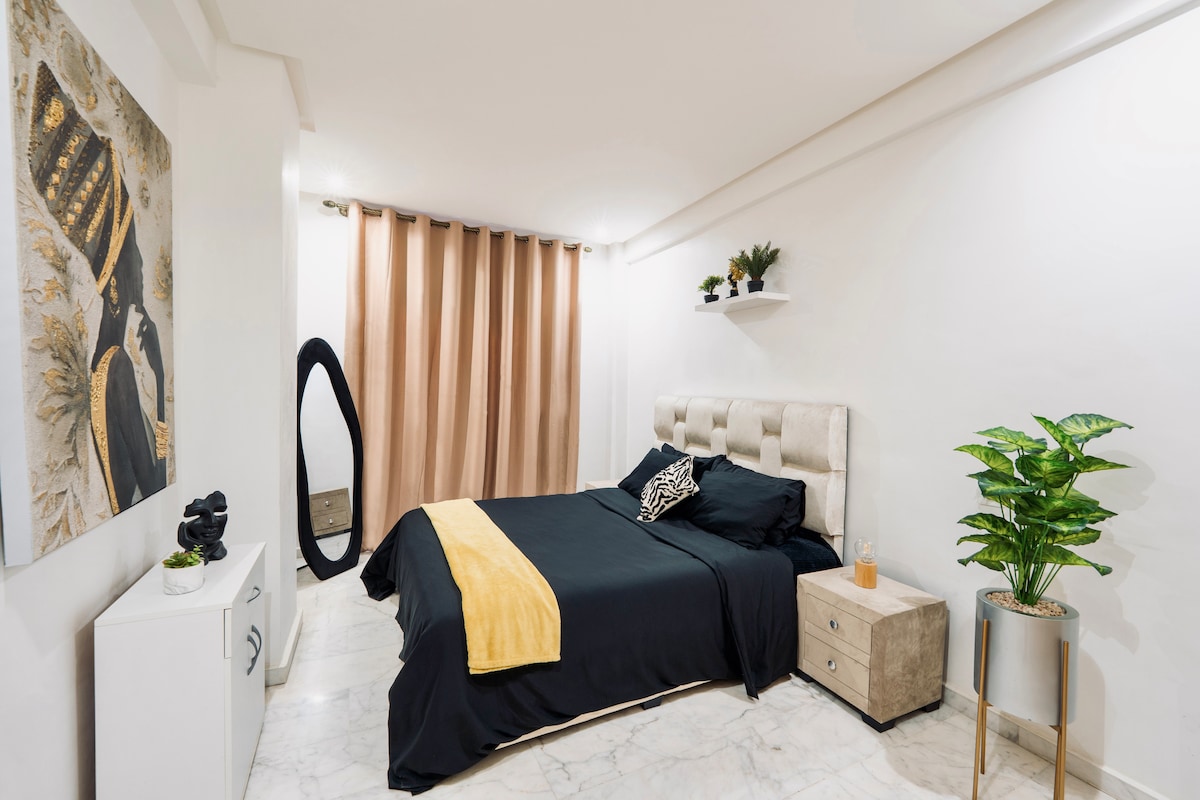 The bedroom showcases a queen-size bed with a black duvet and decorative pillows. A modern headboard is complemented by a beautiful artwork on the wall. A tall mirror stands beside the bed, and a green plant adds a touch of nature. Soft curtains frame the window.