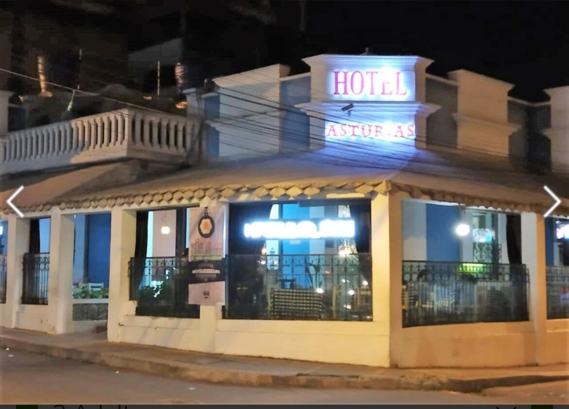 The hotel building features a traditional architectural style, prominently displaying the name 'HOTEL ASTUR' at the top. Large windows are visible, showcasing the interior lighting. A covered balcony adds depth, and the exterior is warmly lit against the night sky.