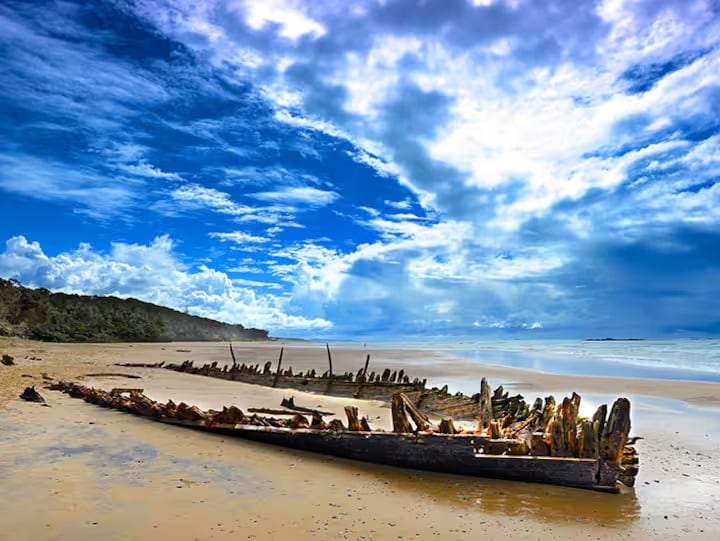 A weathered shipwreck rests on a sandy beach, its wooden remains partially submerged in the shoreline. The sky is filled with dramatic clouds, creating a striking contrast against the calm ocean waters. Lush greenery can be seen in the background along the coastline.