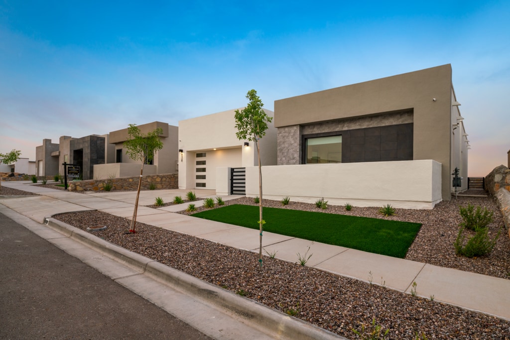 A modern neighborhood is depicted, showcasing several newly constructed houses with a minimalist design. Each home features clean lines and neutral colors, complemented by landscaped gravel beds and patches of grass. Clear skies are visible above, enhancing the serene atmosphere.