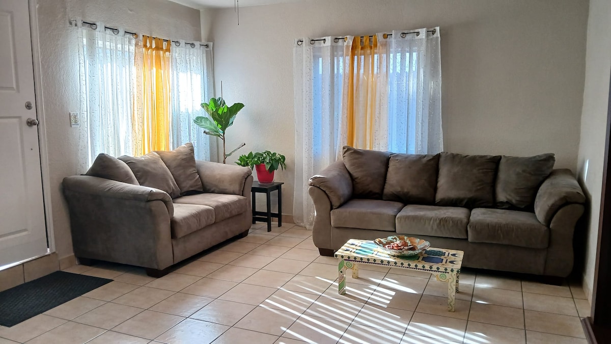 A cozy living area is shown with two plush sofas arranged invitingly. Natural light filters through sheer curtains with vibrant orange accents, highlighting a potted plant situated in a corner. A decorative table holds a colorful tray, adding a touch of charm to the space.