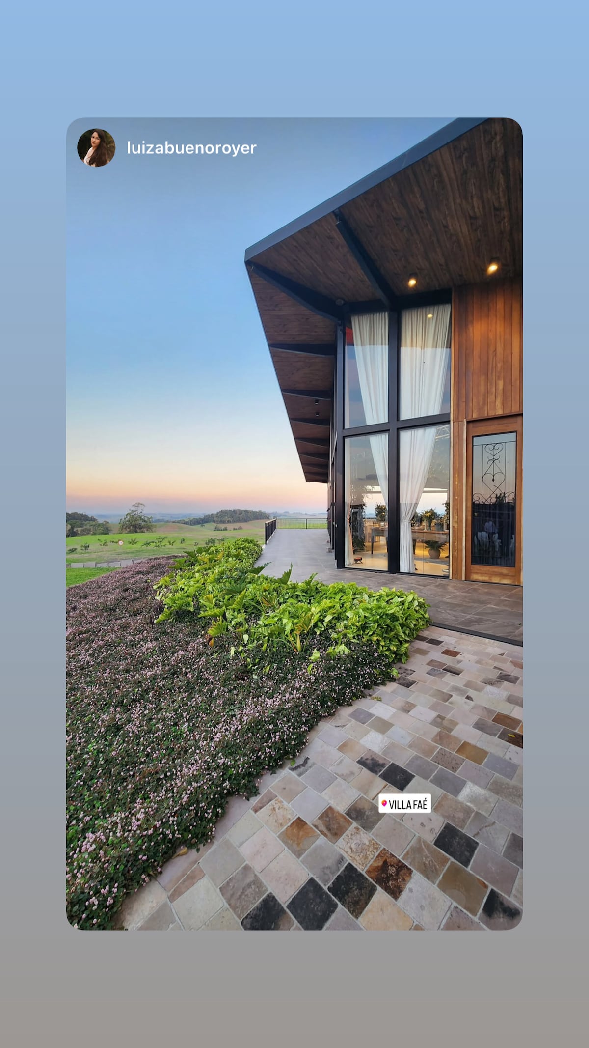 A modern building showcases a wooden exterior, with large glass windows reflecting the colors of the sky. The landscaped area features green shrubbery and a textured stone pathway leading up to the structure, which harmonizes with the natural surroundings.