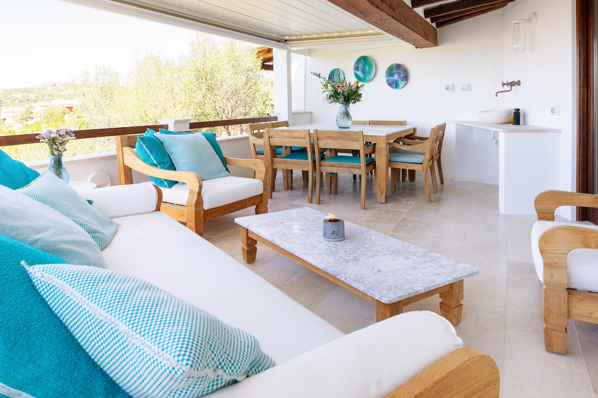 A spacious terrace is furnished with a marble coffee table and a large couch adorned with turquoise and white cushions. A wooden dining table with matching chairs is positioned nearby, complemented by decorative vases. Natural light fills the area, enhancing the inviting space.