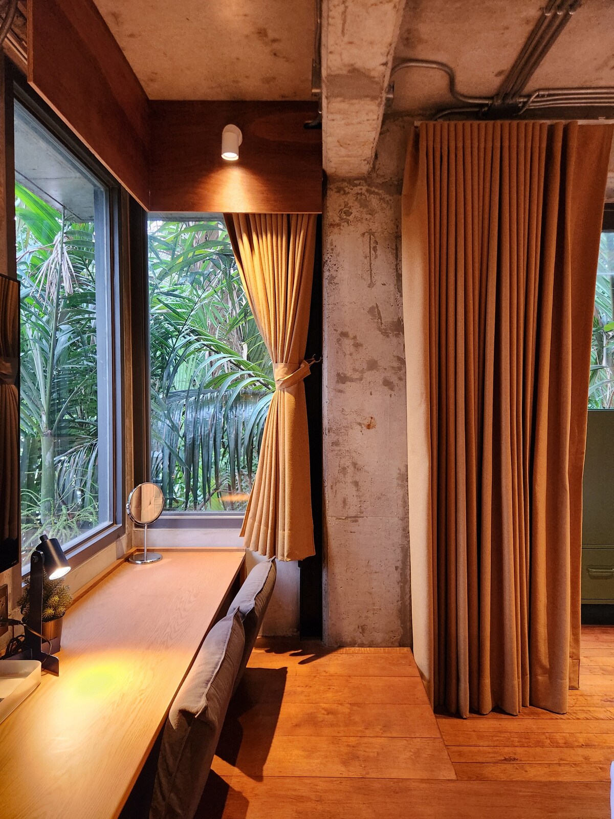 A workspace is situated next to large windows, allowing natural light to enter. Light-colored curtains frame the windows, while a wooden desk and a comfortable chair are arranged neatly. A small mirror and task lamp add functionality to the space, with greenery visible outside.