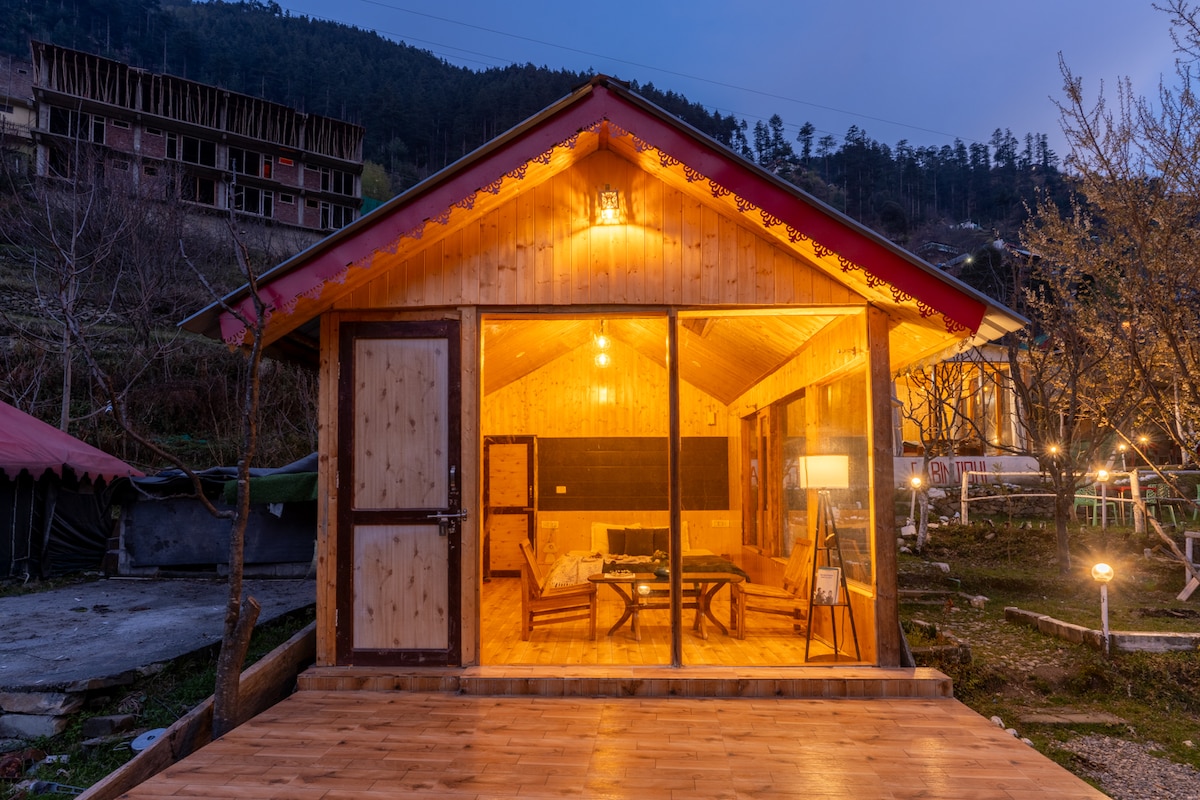 A charming wooden cottage is illuminated by warm lights, creating a welcoming glow. Large glass windows reveal a cozy interior with a seating area and a small dining space. The surrounding nature features trees and the mountainous backdrop, offering a serene atmosphere.
