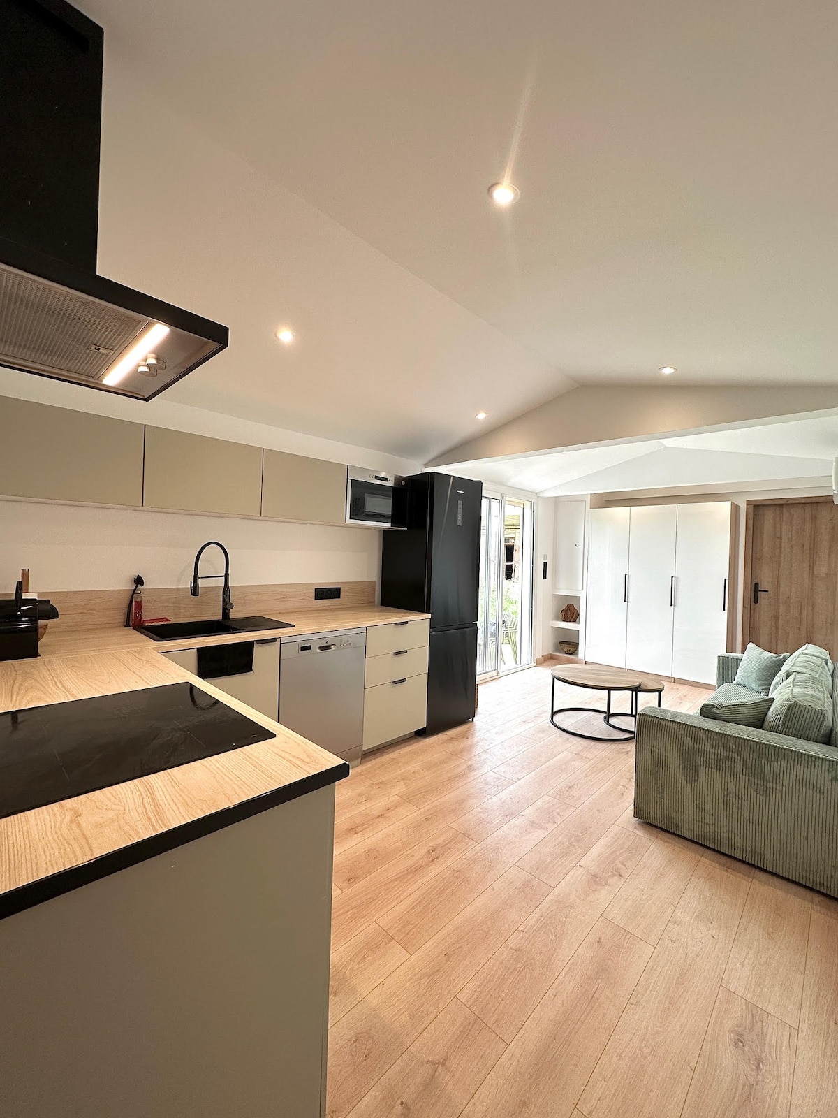 A modern kitchen and living area is depicted, featuring sleek cabinetry and appliances. A countertop with a sink and cooktop is visible, alongside a dining table. Natural light enters from large glass doors leading to an outdoor terrace. A cozy sofa is positioned nearby.