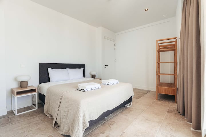 Dieses luftige und minimalistische Schlafzimmer bietet einen ruhigen Rückzugsort mit natürlichen Texturen, warmen Tönen und durchdachter Einfachheit, die zu Ruhe und Entspannung in einem sonnendurchfluteten, übersichtlichen Raum einladen.
