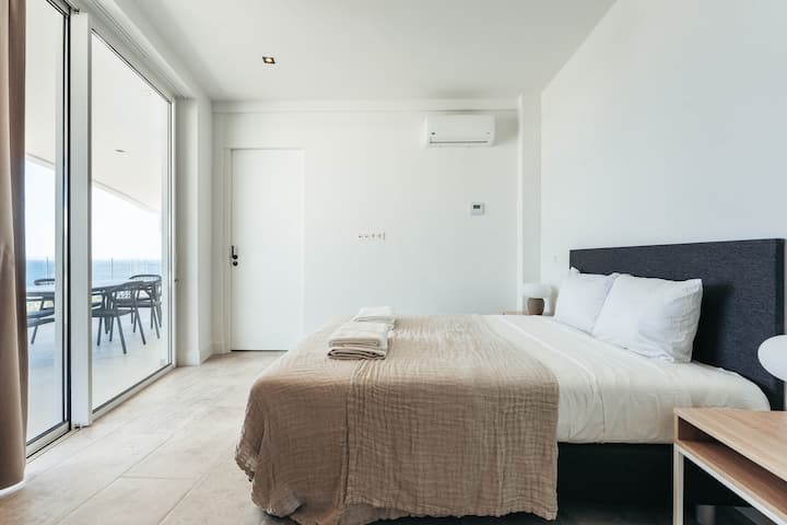 Dieses ruhige Schlafzimmer bietet minimalistischen Luxus – klare Linien, weiche, neutrale Farben und natürliches Licht, das von der Terrasse hereinkommt, schaffen einen ruhigen Rückzugsort, der so beruhigend ist wie die Meeresbrise draußen.