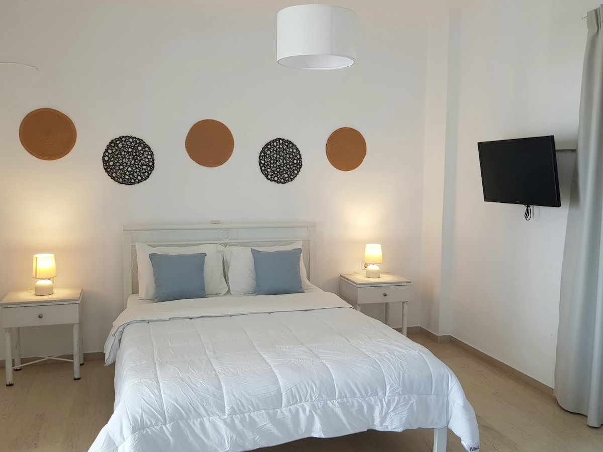 A bright bedroom features a comfortable double bed with soft pillows and a white quilt. Two bedside lamps provide a warm glow on either side. Decorative wall pieces in natural tones complement the neutral color scheme. A wall-mounted television is positioned for easy viewing.