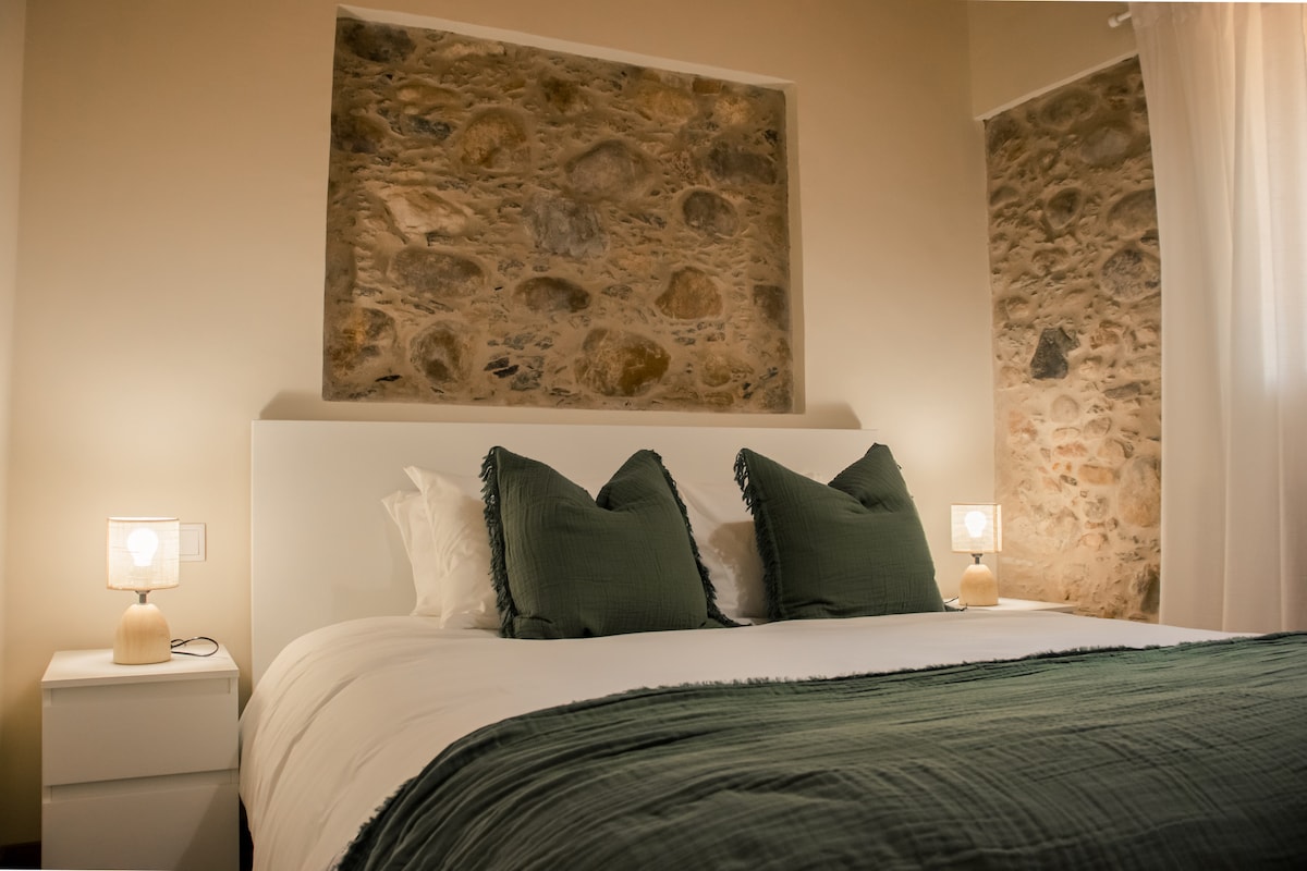 A bedroom features a spacious bed with textured green pillows, positioned against a rustic stone wall. Soft bedside lamps provide gentle illumination on either side of the bed, while light drapes frame a window, enhancing the serene atmosphere.