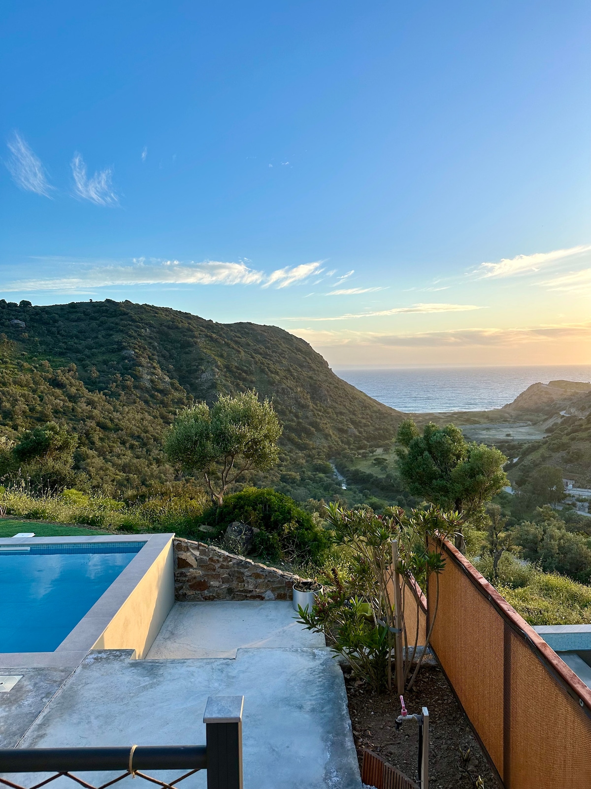 A view of the private swimming pool is captured, with rugged hills and the sea visible in the background. The sun sets behind the landscape, casting warm hues across the sky. Lush greenery and a stone wall frame the serene outdoor space.