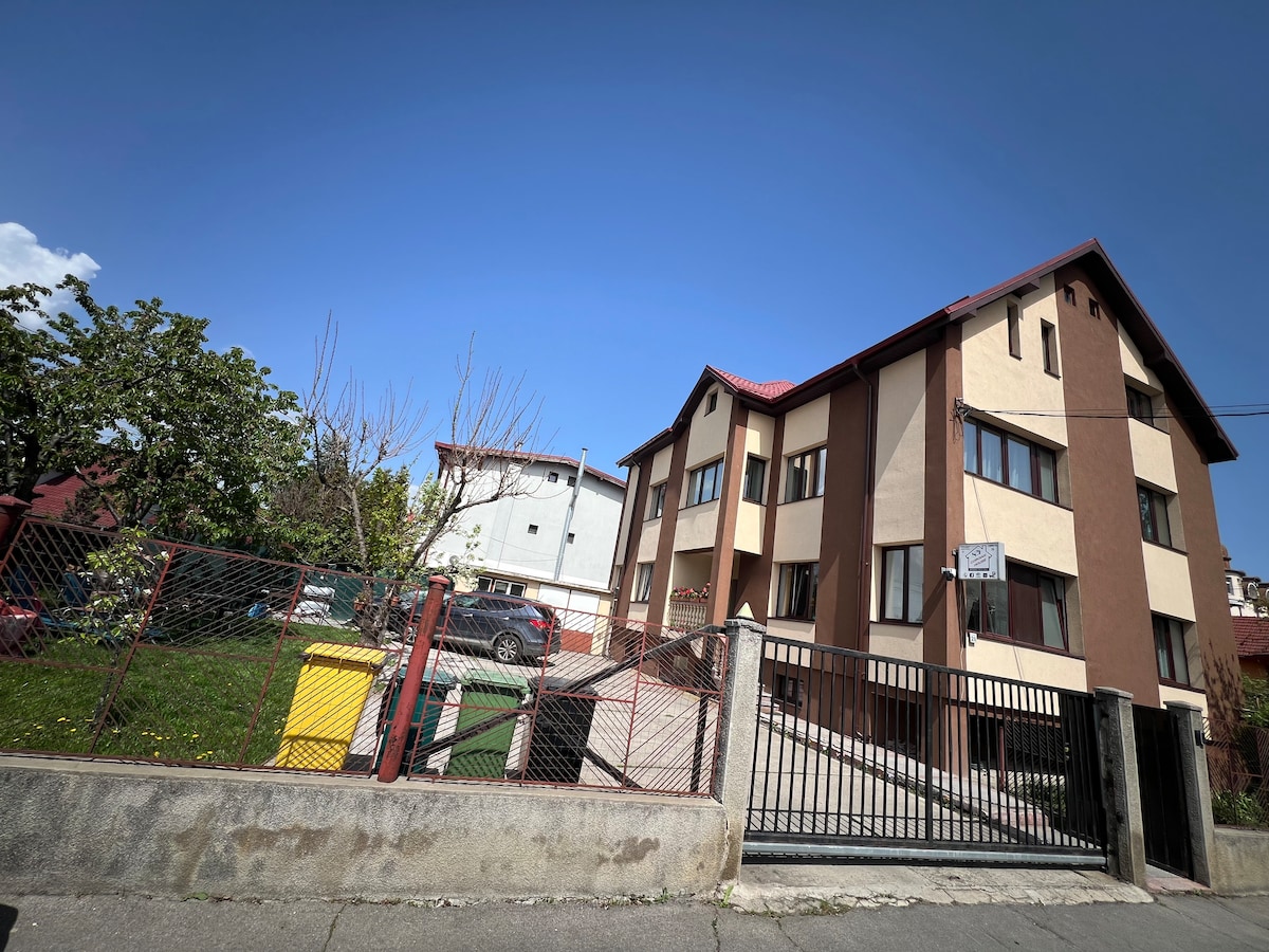 A multi-story building is depicted with a mixture of brown and beige facade. The entrance features a gated pathway leading to the front door, surrounded by a landscaped yard with trees. Parking spaces are visible at the side, offering private parking options.