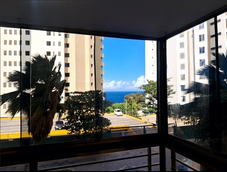 A view from a large window showcases the ocean in the distance, framed by palm trees and surrounding buildings. Bright blue skies and fluffy white clouds enhance the coastal scenery, while natural light fills the interior space.