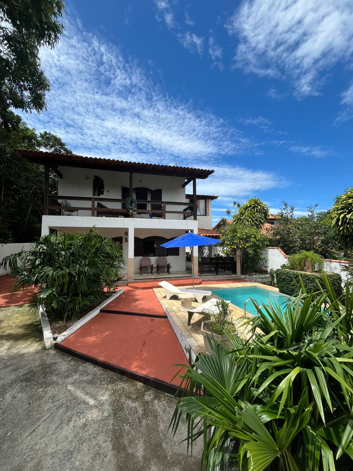 A spacious house is set against a vibrant blue sky, featuring a terrace overlooking a pool. Lush greenery frames the area, with a sun umbrella positioned near lounge chairs beside the water. The building displays a traditional architectural style with multiple balconies.