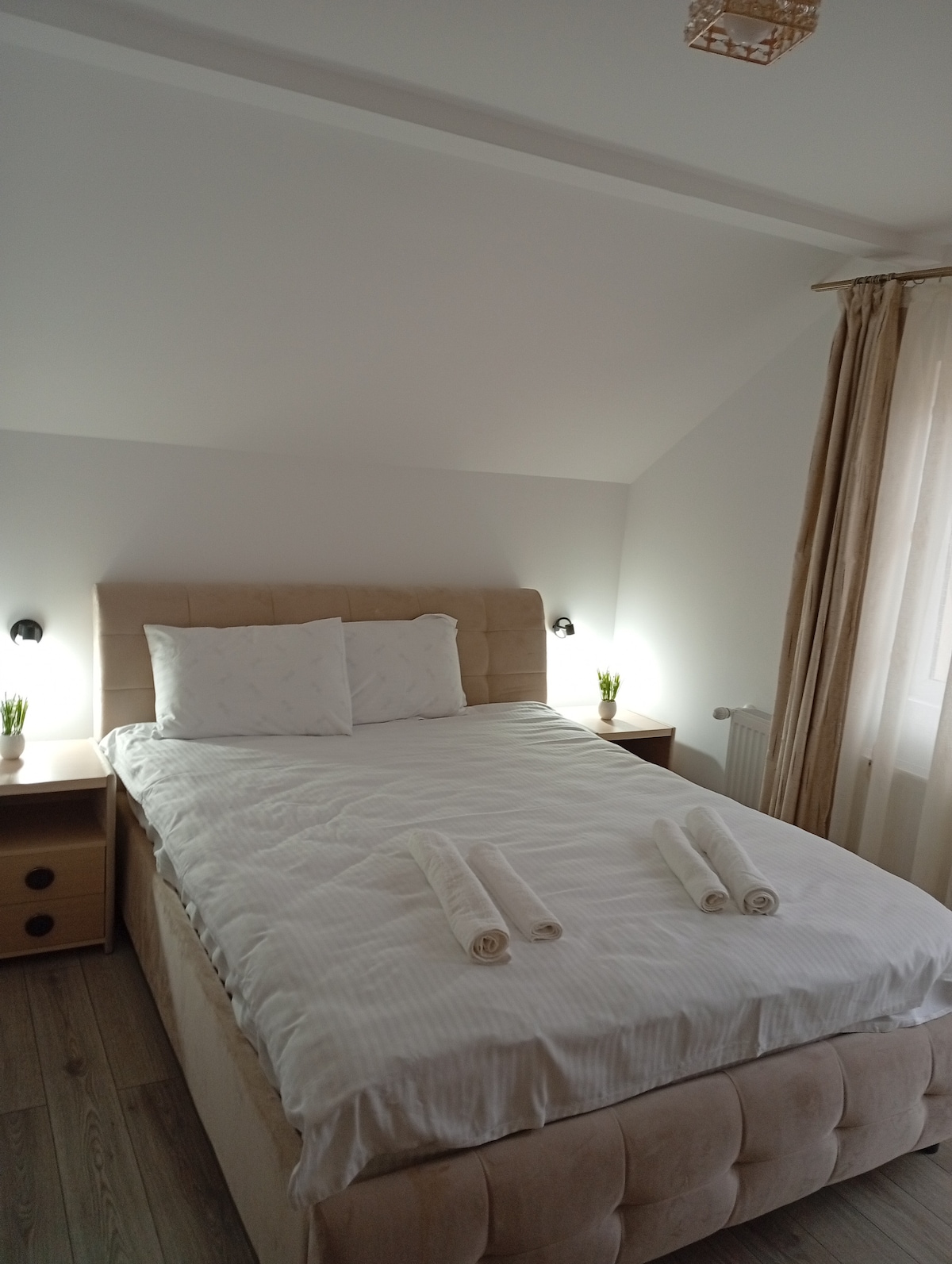 A well-lit bedroom features a spacious bed dressed in crisp white linens. Two rolled towels are placed neatly at the foot of the bed. Nightstands are positioned on either side, each with small potted plants. Light filters softly through sheer curtains, enhancing the serene atmosphere.