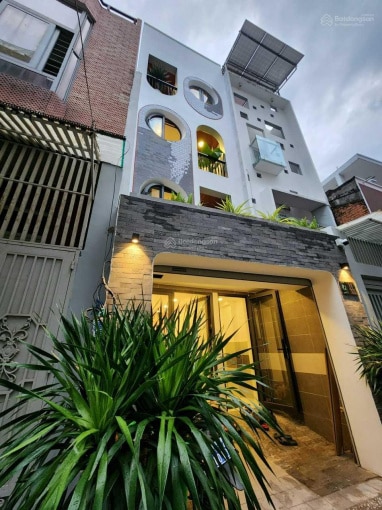 The exterior of a modern building is showcased, featuring a blend of smooth and textured surfaces. Lush greenery lines the entrance, complemented by round windows and a balcony above. Soft lighting illuminates the entryway, enhancing the welcoming appearance of the structure.