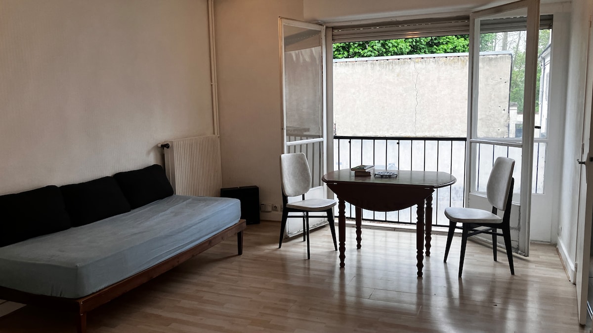 A spacious living area is illuminated by natural light streaming through large glass doors. A pull-out sofa bed is positioned beside a round wooden table, accompanied by two upholstered chairs. The area offers a view of a small balcony, enhancing the open feel.